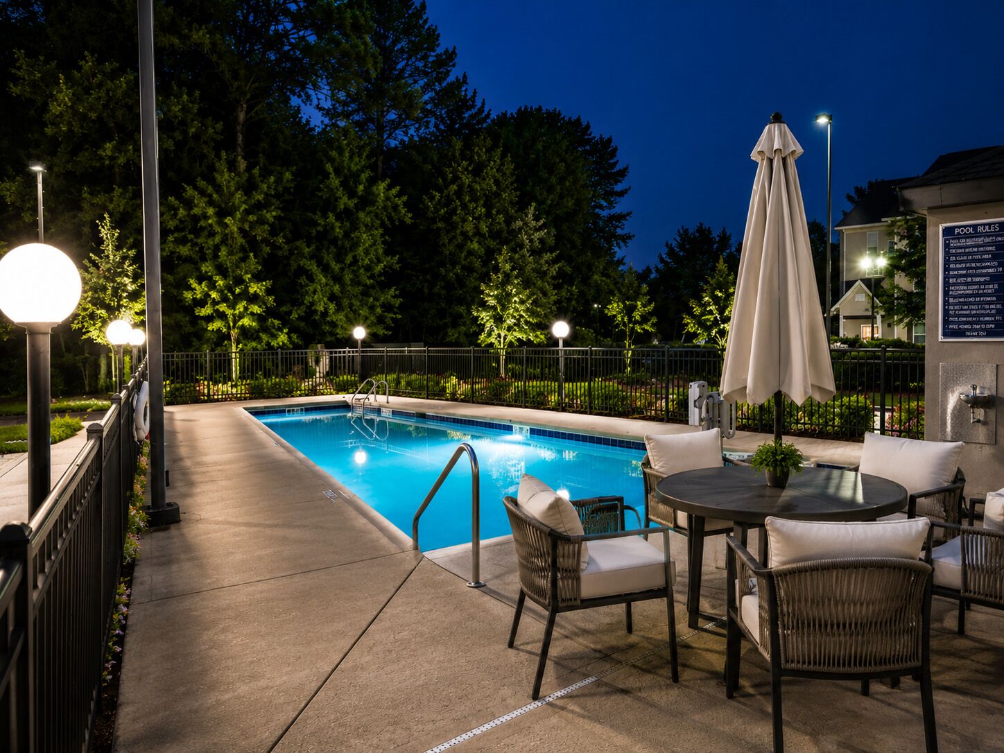 A swimming pool with a paved deck, surrounded by a fence, and outdoor seating with a table and chairs.
