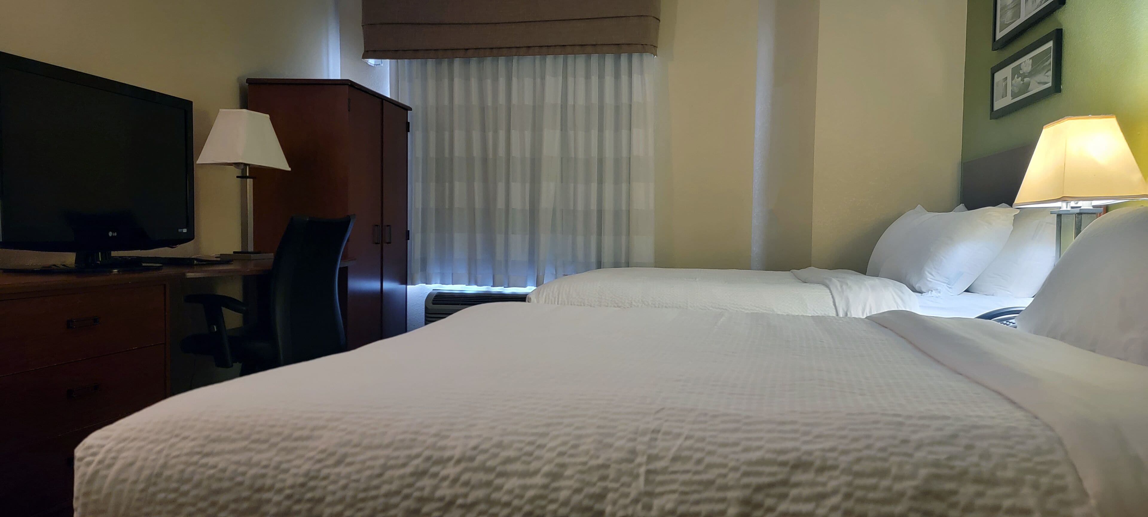 A hotel room with a bed, a desk with a TV, a chair, a wardrobe, and a window with curtains.
