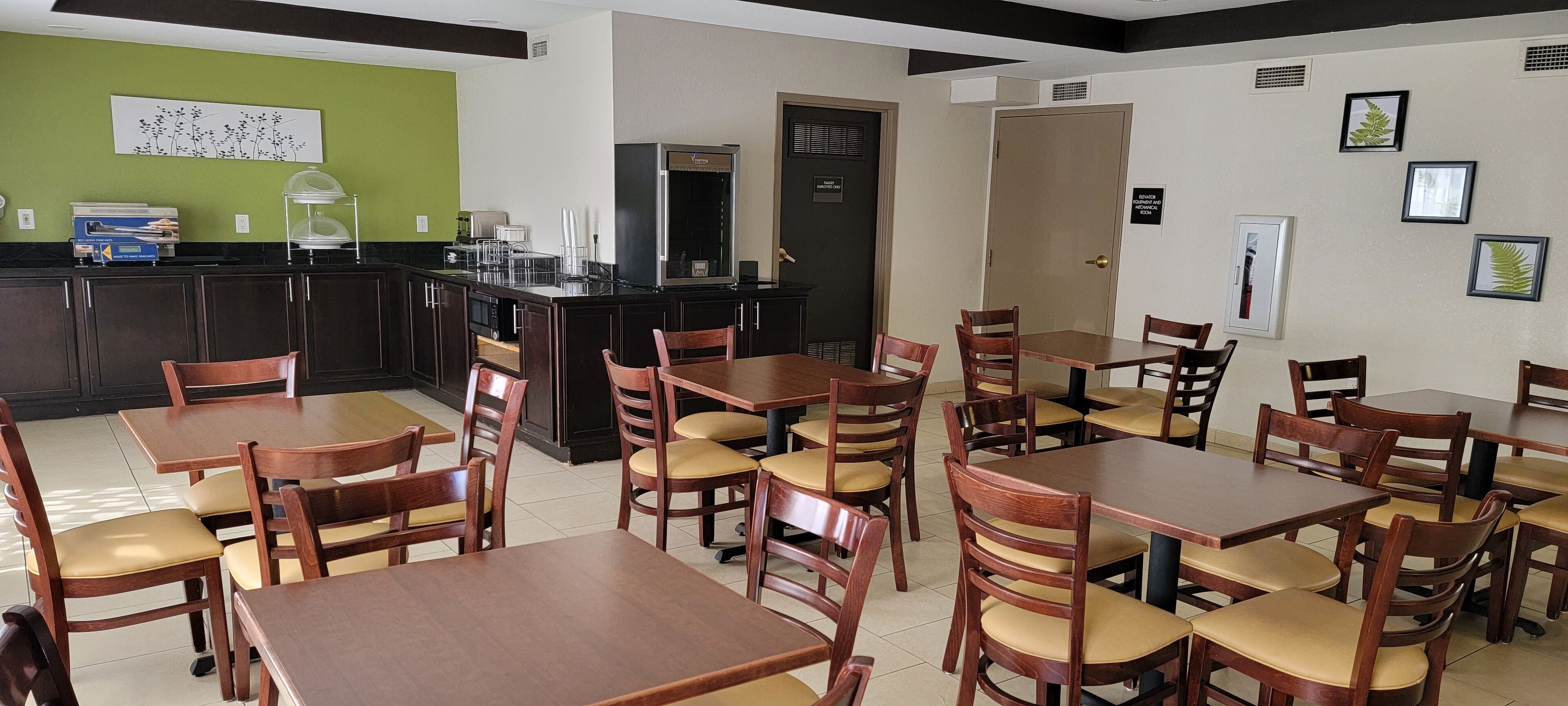 A dining area with wooden tables and chairs, a kitchen area with a refrigerator, and a green wall with a floral artwork.