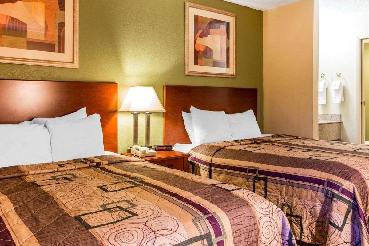 A hotel room with two beds, a nightstand, a lamp, and two framed abstract paintings on the walls.
