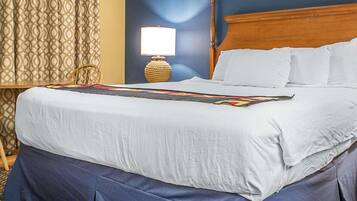 Una cama bien hecha con una colcha blanca y falda azul, cabecera de madera, lámpara de noche y cortina estampada.