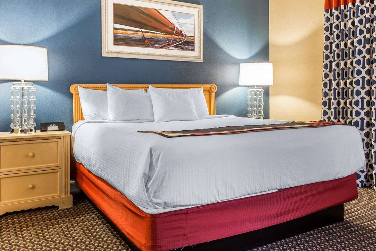 Une chambre d’hôtel avec un lit, deux tables de chevet, une petite table, un tableau encadré au mur et un rideau à motifs.