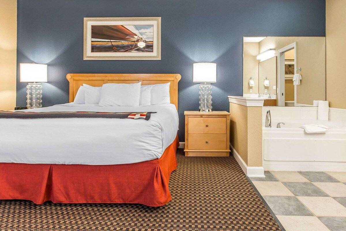 Une chambre d’hôtel comprenant un lit, des lampes de chevet, une table de chevet, une salle de bain avec un lavabo et un miroir, et un tapis à motifs.