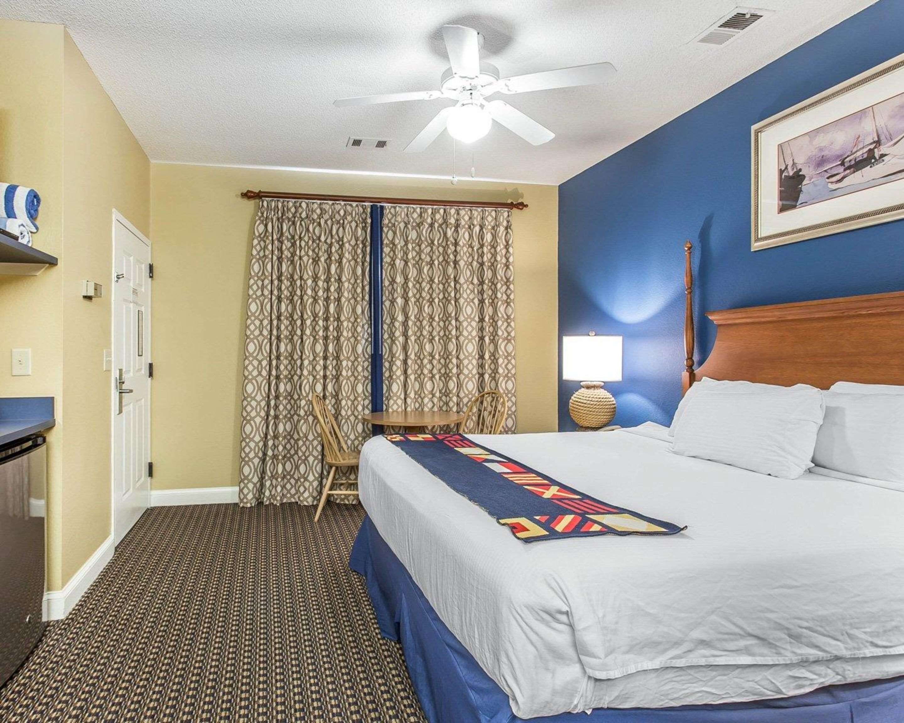 Une chambre d’hôtel équipée d’un lit, d’un bureau, d’une chaise, d’un ventilateur de plafond, de rideaux et d’une lampe fixée au mur.