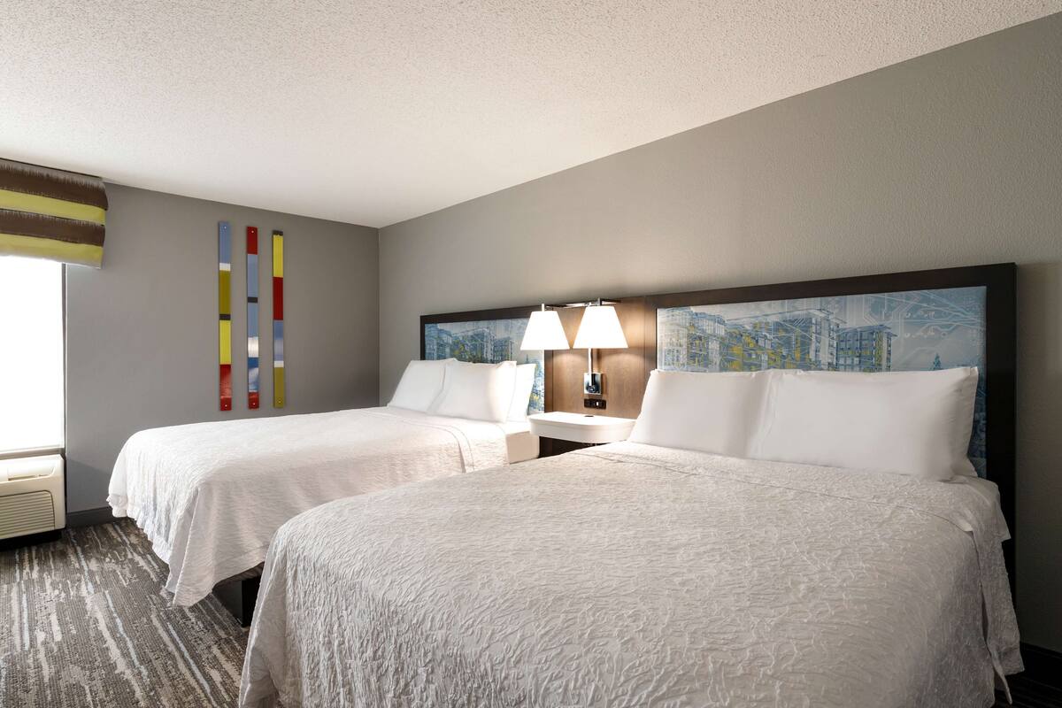 A hotel room with two beds, a cityscape painting on the headboard, a lamp, and a window with blinds.