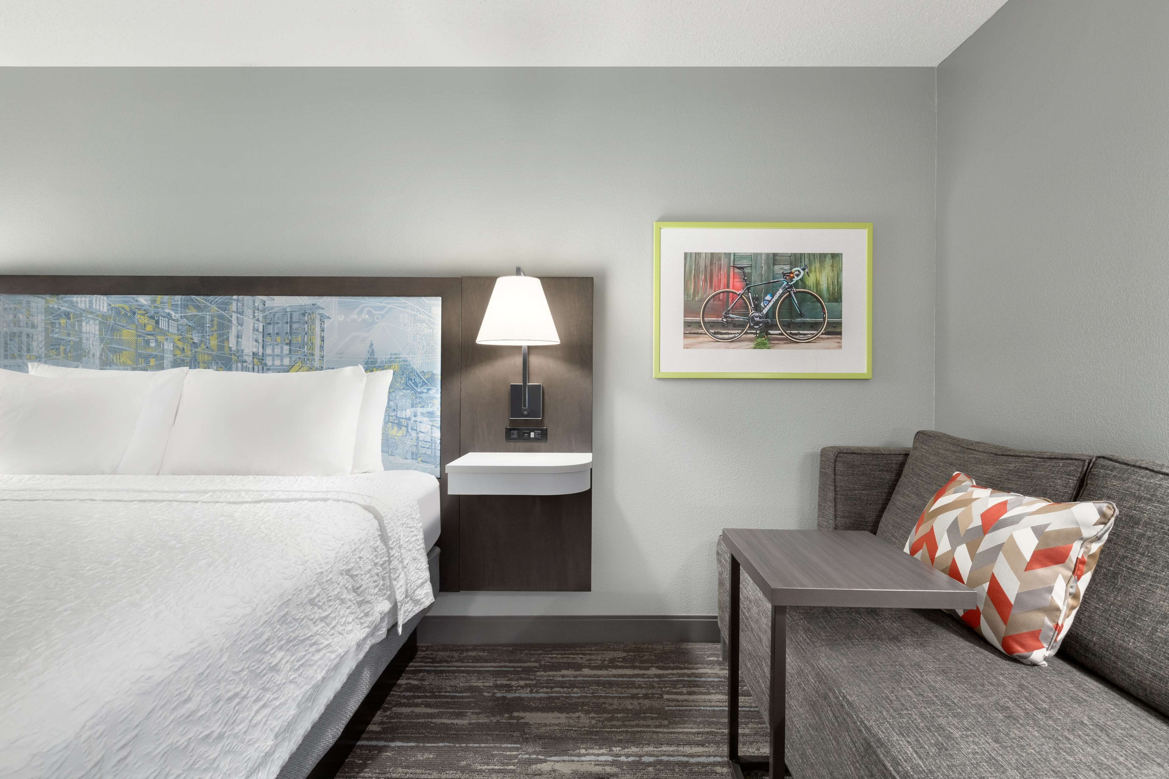 A hotel room with a bed, a nightstand with a lamp, a small table, a sofa with a patterned pillow, and a framed picture on the wall.