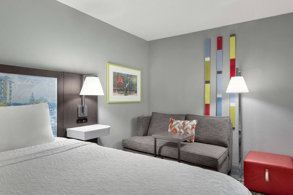 A hotel room with a bed, a sofa, a small table, a red suitcase, a lamp, a picture frame, and a wall with vertical stripes.