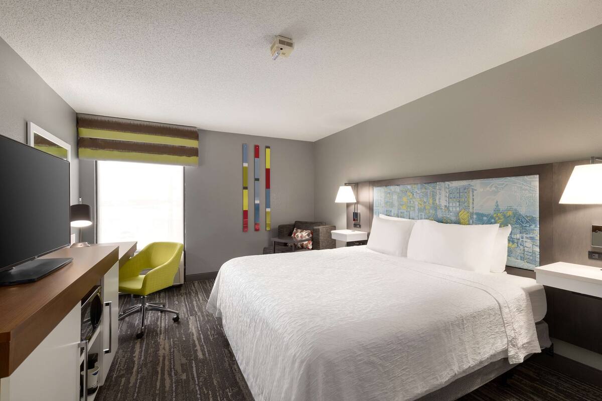 A hotel room with a large bed, a desk with a computer, a chair, a desk lamp, a nightstand, and a painting on the wall.