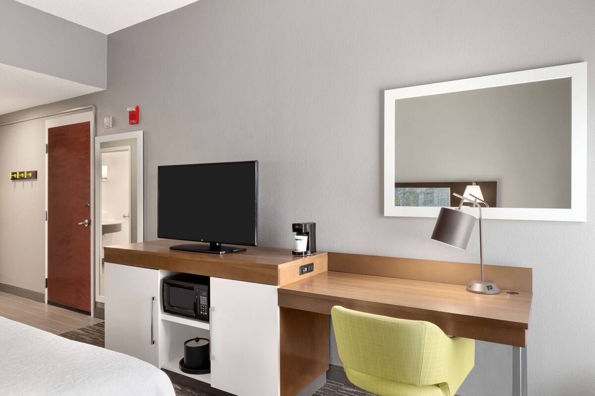 A hotel room with a wooden desk, a television, a coffee maker, a mirror, and a green chair.