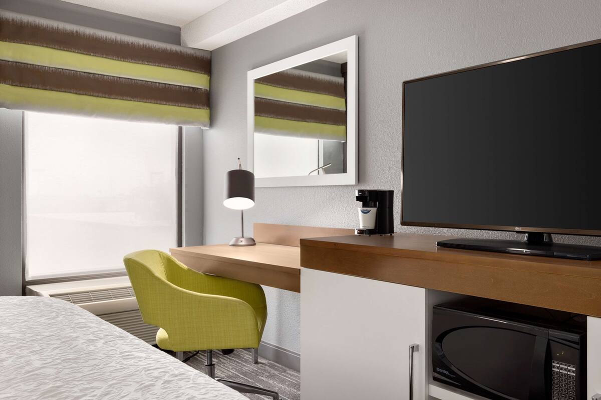 A hotel room with a bed, a desk with a chair, a television, a mirror, and a window with curtains.