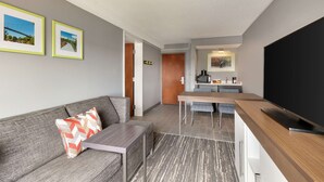 A modern hotel room with a grey sofa, a small table, a TV, and a kitchen area with a microwave and a painting. - Hampton Inn Simpsonville (Simpsonville)