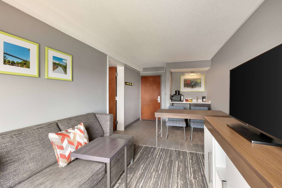 A modern hotel room with a grey sofa, a small table, a TV, and a kitchen area with a microwave and a painting.