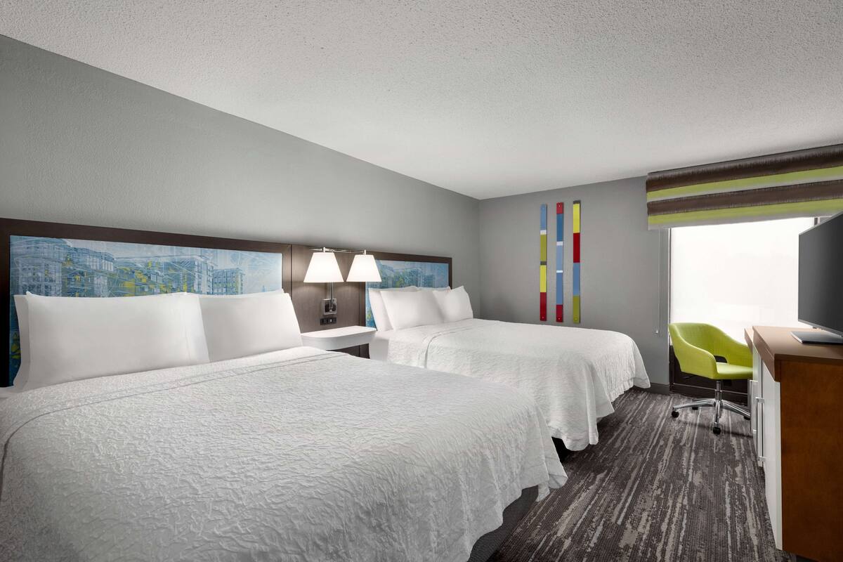 A hotel room with two beds, a desk, a chair, and a cityscape painting on the wall.