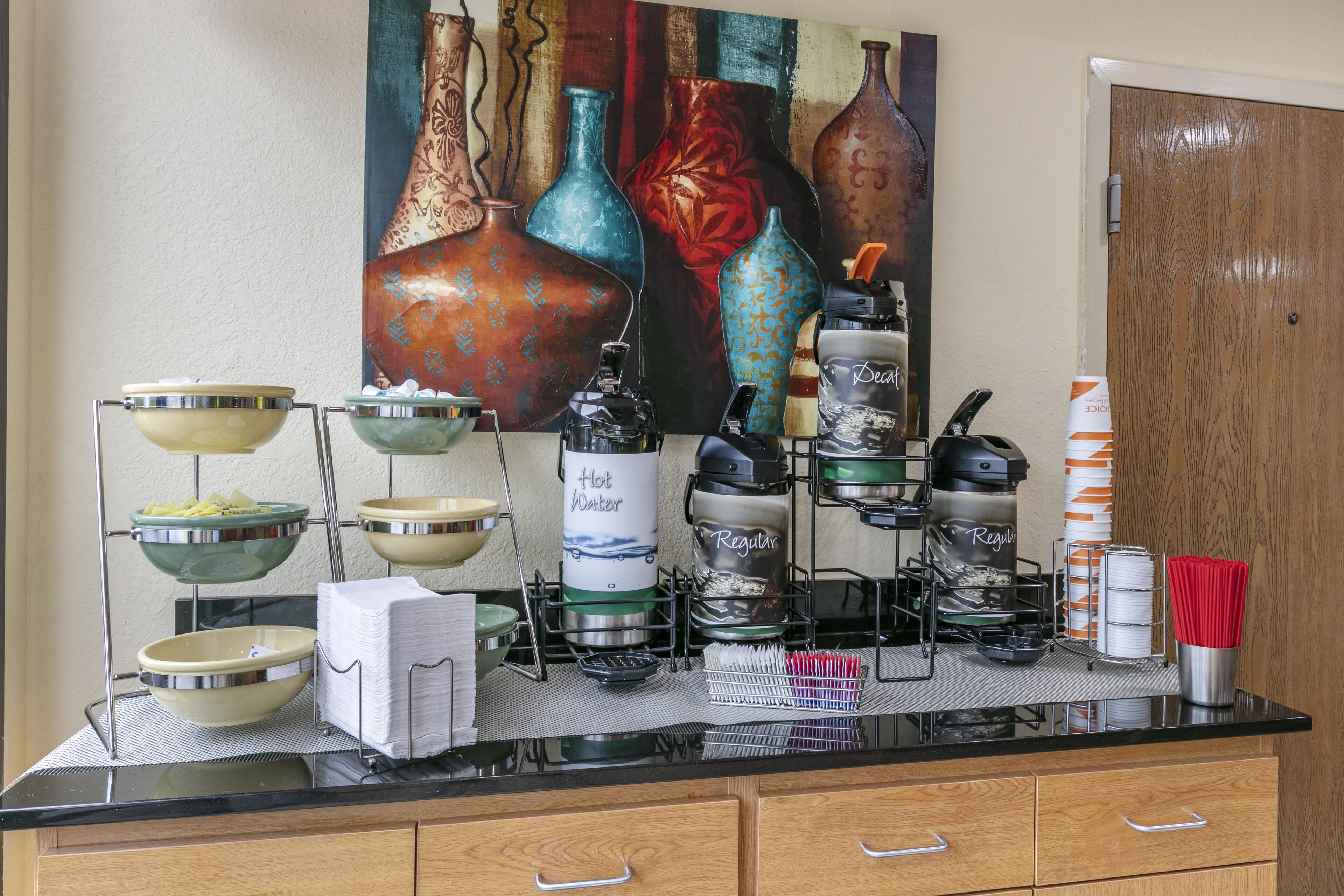 Un buffet composé de différents plats et d’ustensiles de service, une machine à café et un tableau décoratif représentant des vases accroché au mur.