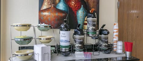 Un buffet composé de différents plats et d’ustensiles de service, une machine à café et un tableau décoratif représentant des vases accroché au mur.