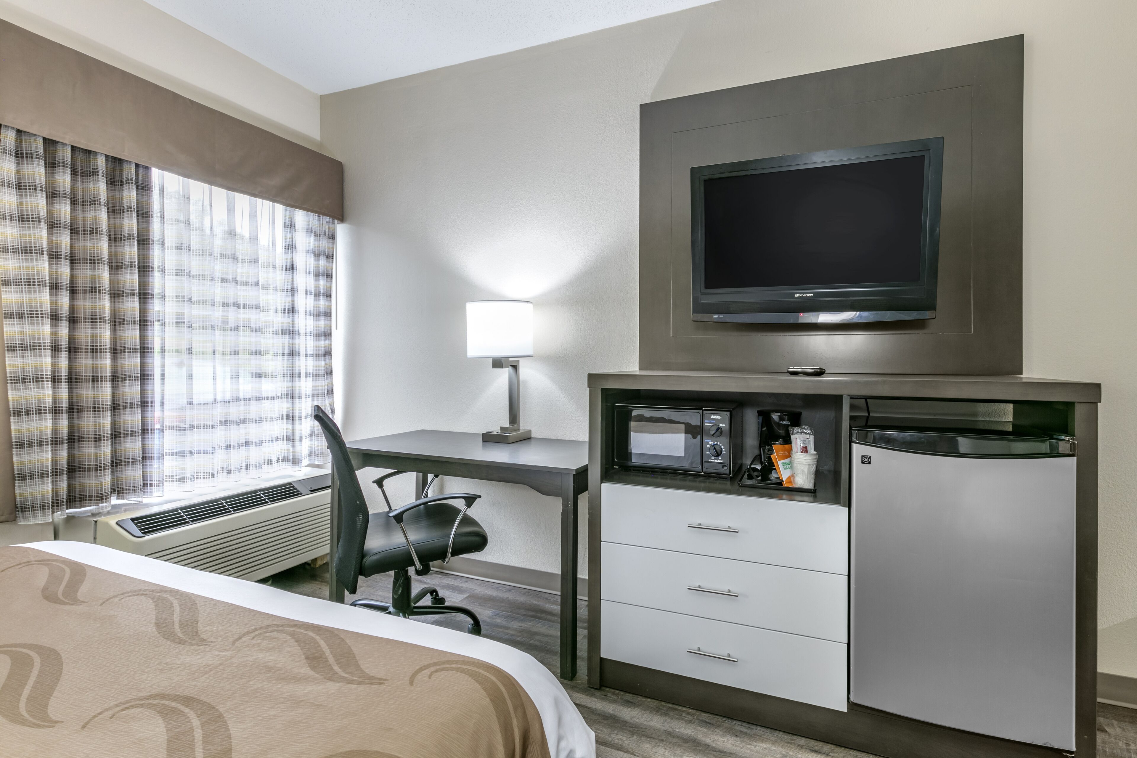 A hotel room with a bed, desk, chair, television, microwave, refrigerator, and a window with curtains.