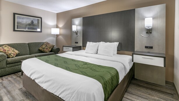 A hotel room with a large bed, a green sofa, a nightstand with a lamp, and a wall-mounted light fixture. - Quality Inn Rogersville (Rogersville)