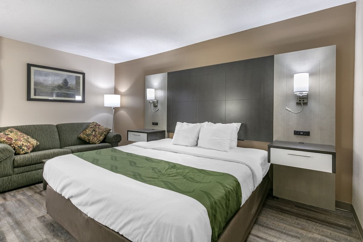 A hotel room with a large bed, a green sofa, a nightstand with a lamp, and a wall-mounted light fixture.