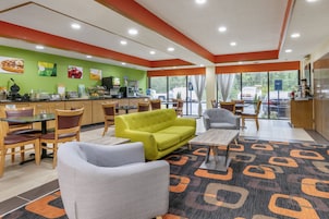 A modern hotel lobby with a green wall, a coffee station, seating areas, and large windows offering a view of the parking lot.