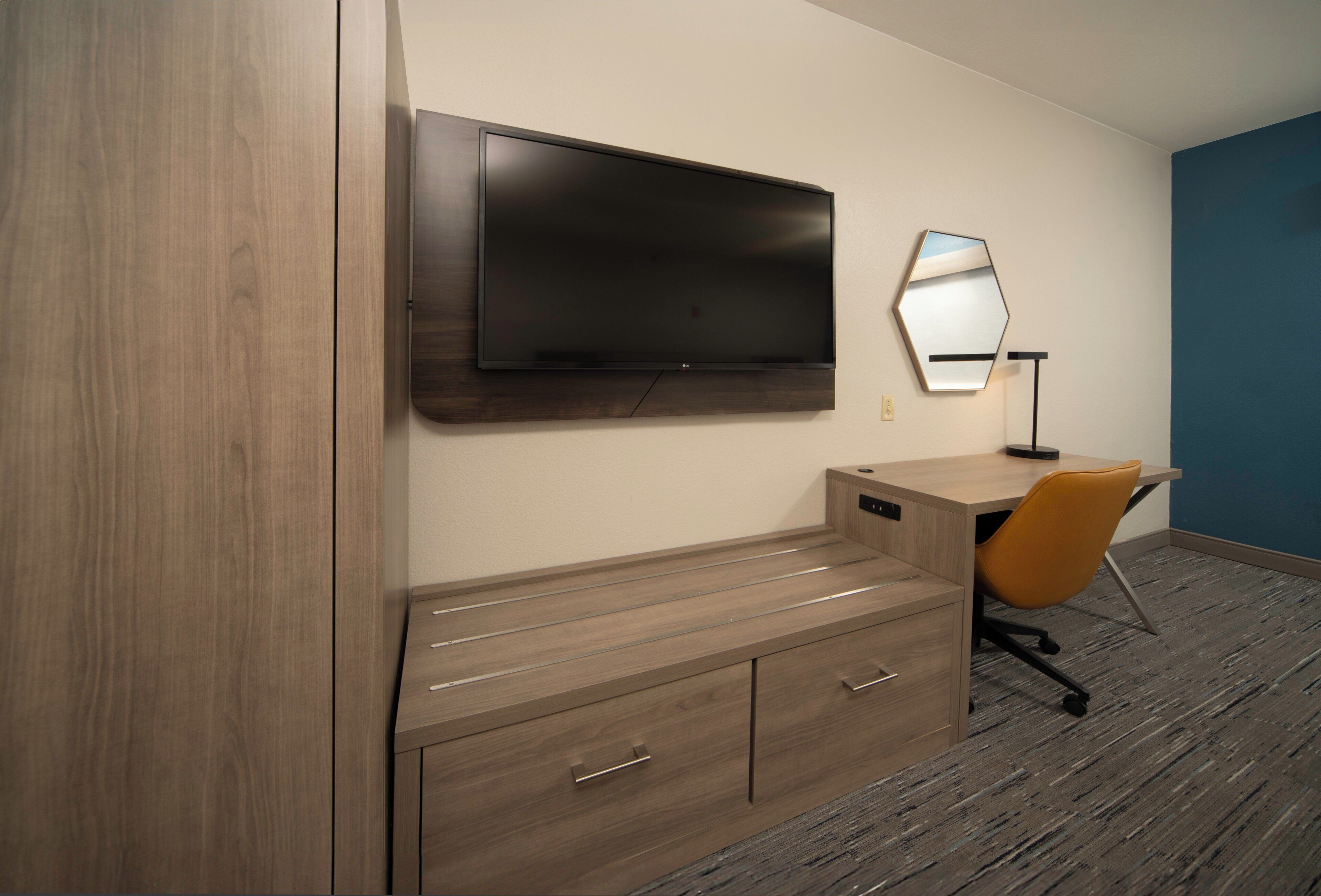 A hotel room with a flat-screen TV mounted on the wall, a wooden desk with drawers, an orange office chair, and a mirror.