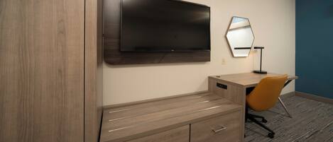A hotel room with a flat-screen TV mounted on the wall, a wooden desk with drawers, an orange office chair, and a mirror.