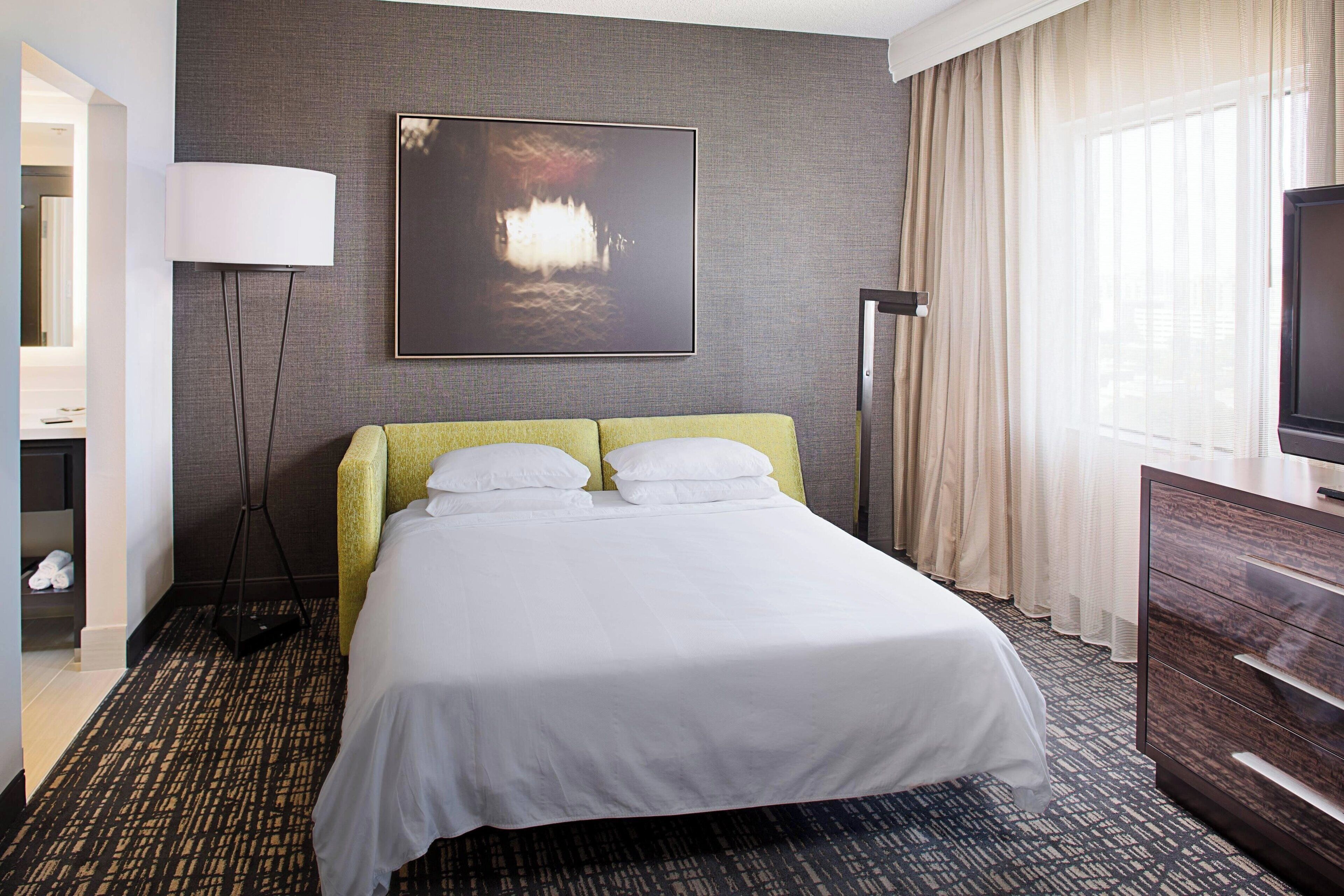 Una habitación de hotel con una cama grande, un televisor de pantalla plana, una lámpara de pie y un cuadro en la pared.