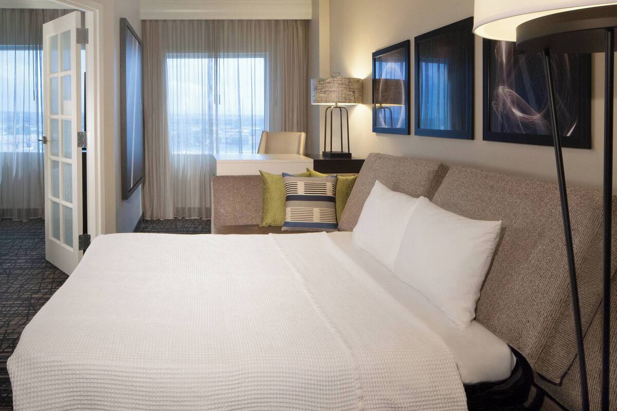 Habitación de hotel con una cama grande, un escritorio con lámpara, una silla y vista a la ciudad por la ventana.