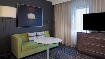 A hotel room with a green sofa, a round table, a flat-screen TV, and a large window with curtains.