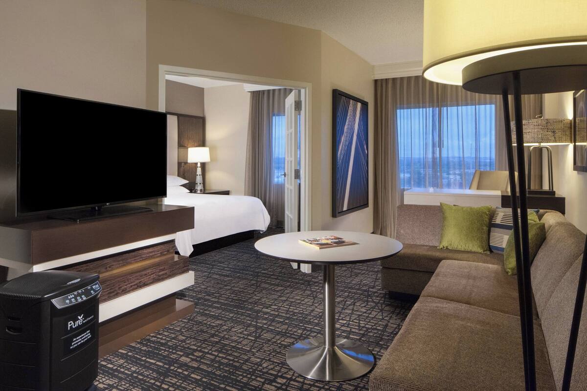 Habitación de hotel con una gran televisión de pantalla plana, una cama con ropa blanca, un sofá, una mesita redonda y una lámpara.