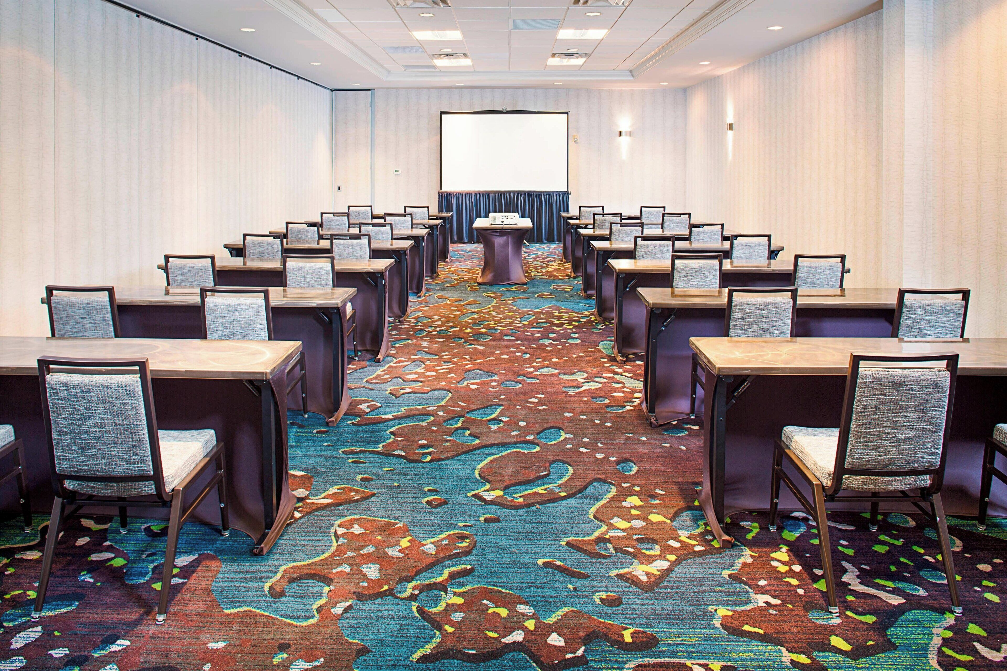 Une salle de conférence avec des rangées de tables et de chaises, un sol recouvert de moquette et un grand écran à l’avant.