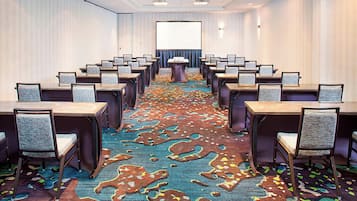 Une salle de conférence avec des rangées de tables et de chaises, un sol recouvert de moquette et un grand écran à l’avant.