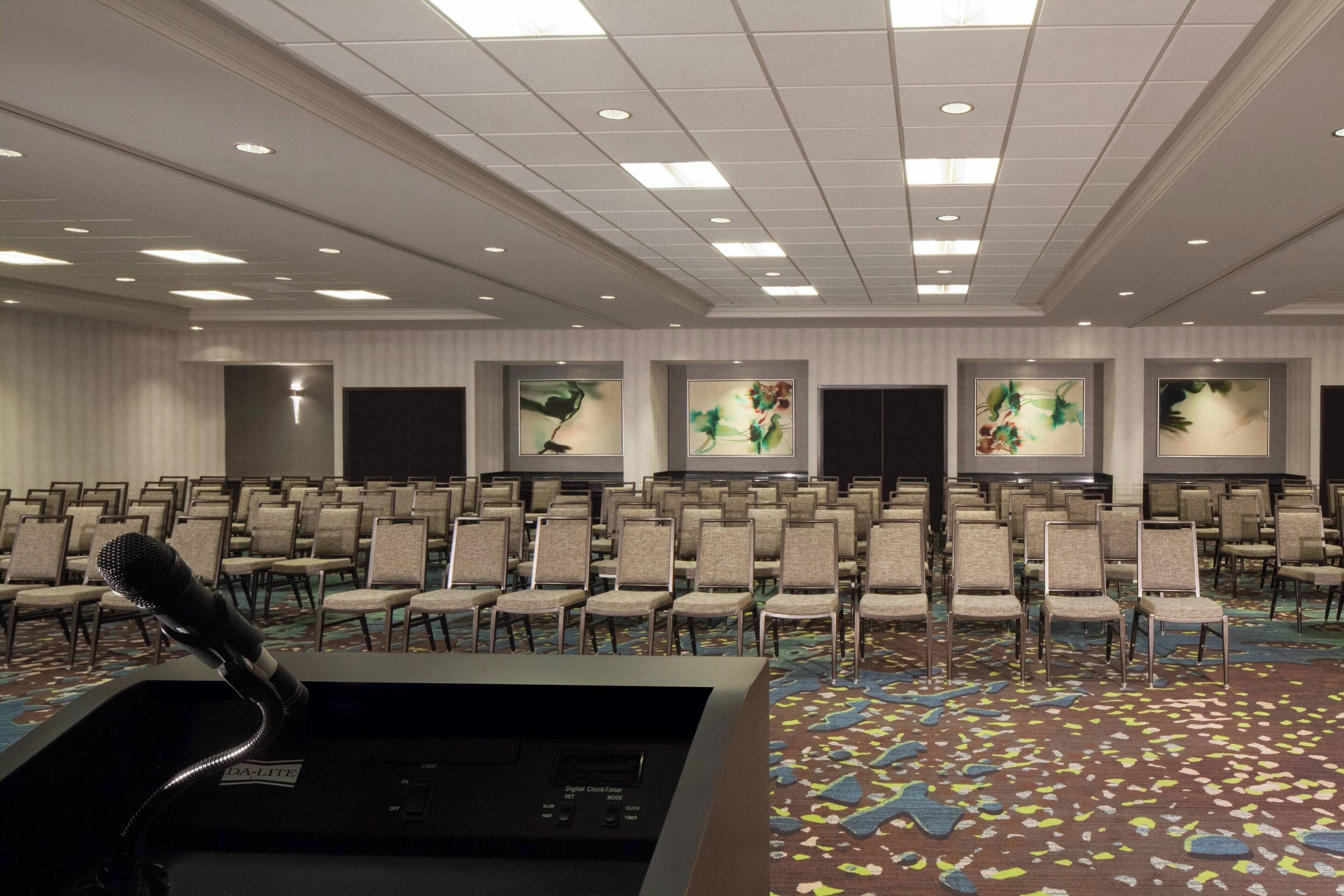 A conference room with rows of chairs, a podium with a microphone, and framed artwork on the walls.