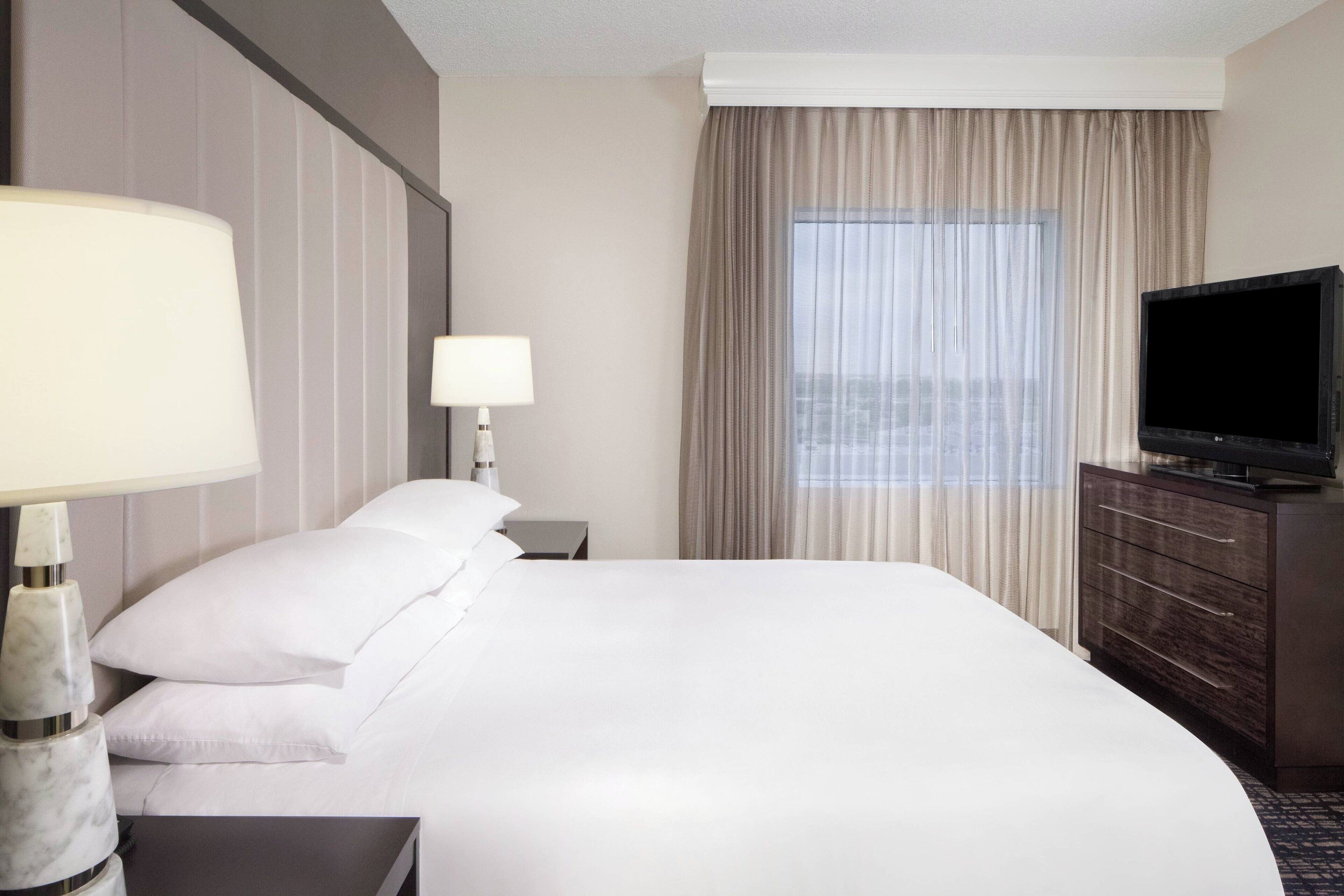 A hotel room with a large bed, a TV on a wooden dresser, a nightstand with a lamp, and a window with curtains.