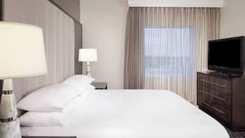 Une chambre d’hôtel avec un grand lit, un téléviseur posé sur une commode en bois, une table de chevet avec une lampe et une fenêtre avec des rideaux.