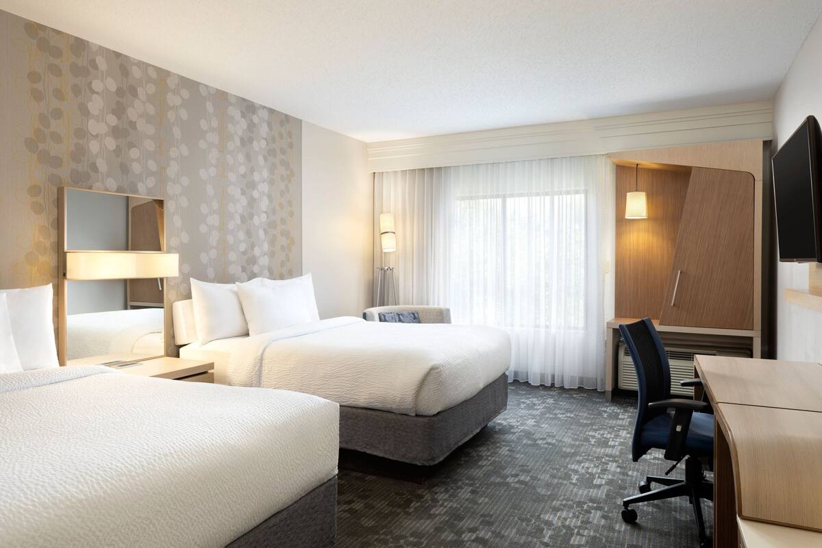 A hotel room with two beds, a desk, a chair, a TV, and a large window with curtains.