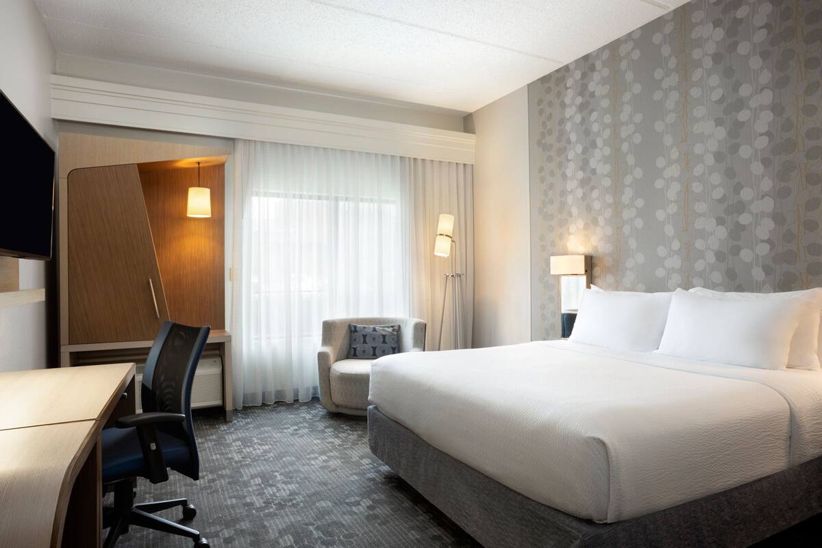 A hotel room with a large bed, a desk, a chair, and a small armchair.