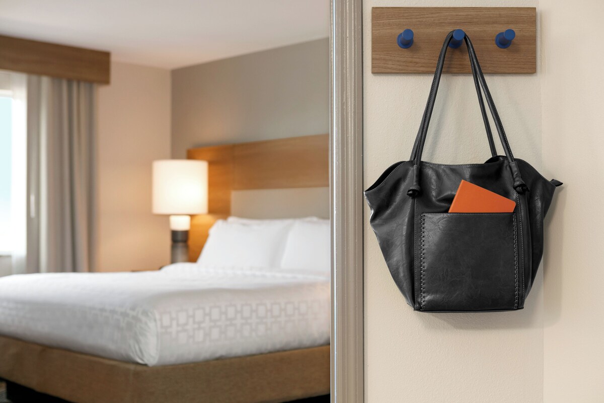 A hotel room with a neatly made bed, a bedside lamp, and a wall-mounted coat hook with a black leather bag.