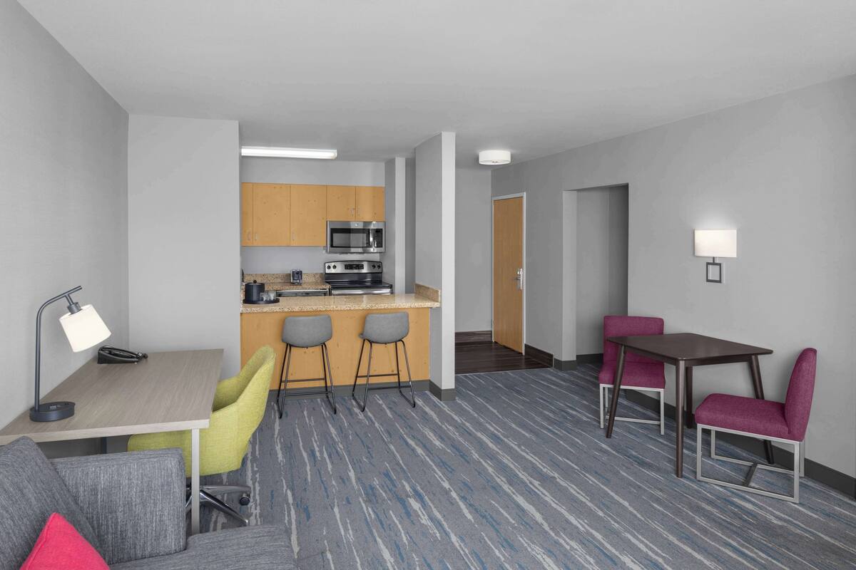 Habitación de hotel con kitchenette, escritorio con silla, mesa de comedor con sillas y piso alfombrado.