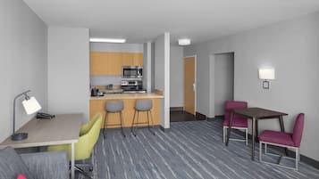 A hotel room with a kitchenette, a desk with a chair, a dining table with chairs, and a carpeted floor.