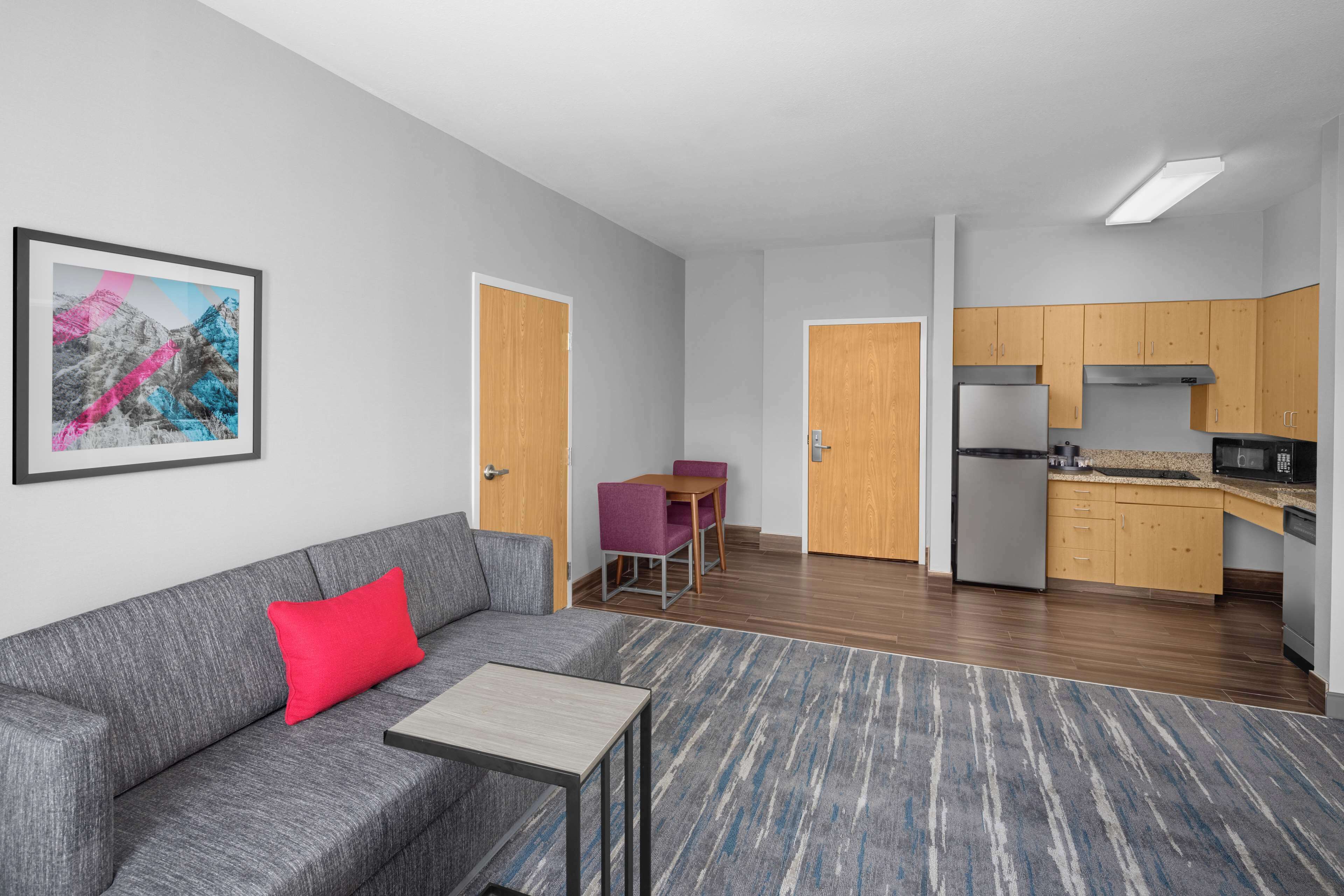 A modern apartment with a grey sofa, a small table, a dining area with a purple chair, and a kitchen with wooden cabinets and a refrigerator.