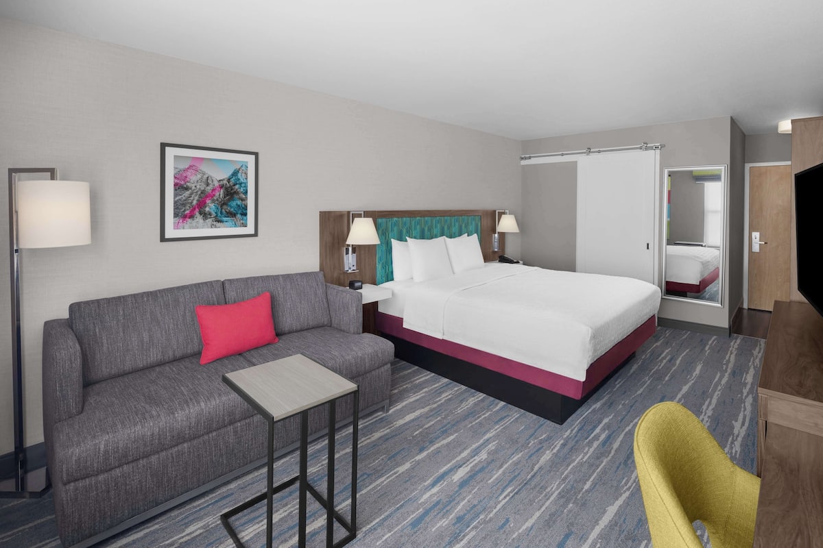 Habitación de hotel con cama, sofá, mesita, silla, lámpara, un cuadro y armario.