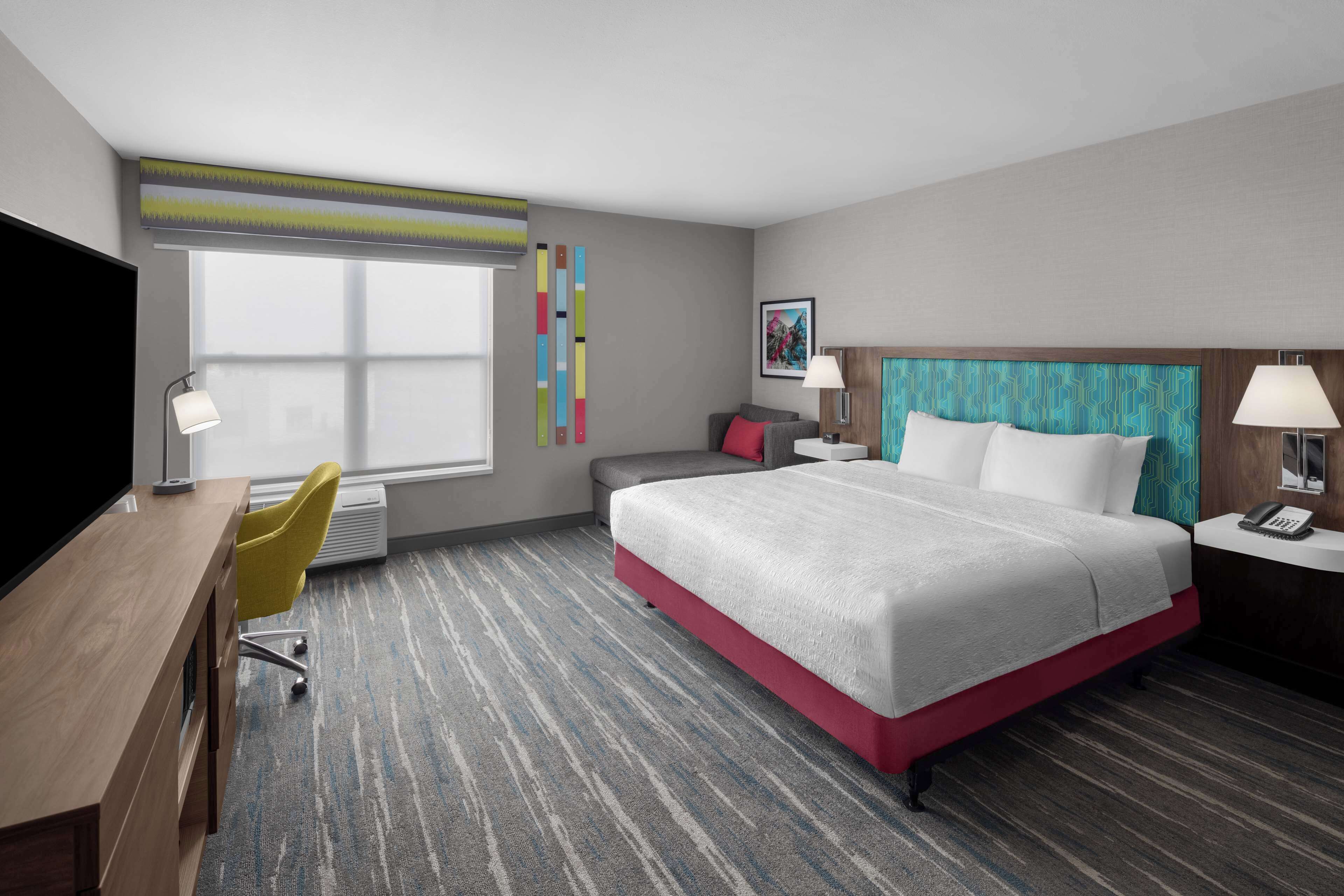 A hotel room with a large bed, a desk, a chair, a TV, and a window with blinds.