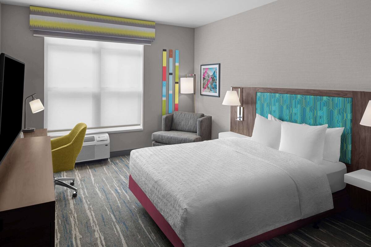 Habitación de hotel con una cama grande, un escritorio con una silla, una ventana con persianas y una pared decorada con una obra de arte colorida.