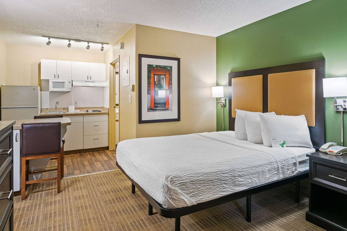 A hotel room with a bed, a desk, a chair, a microwave, a sink, and a framed picture on the wall.