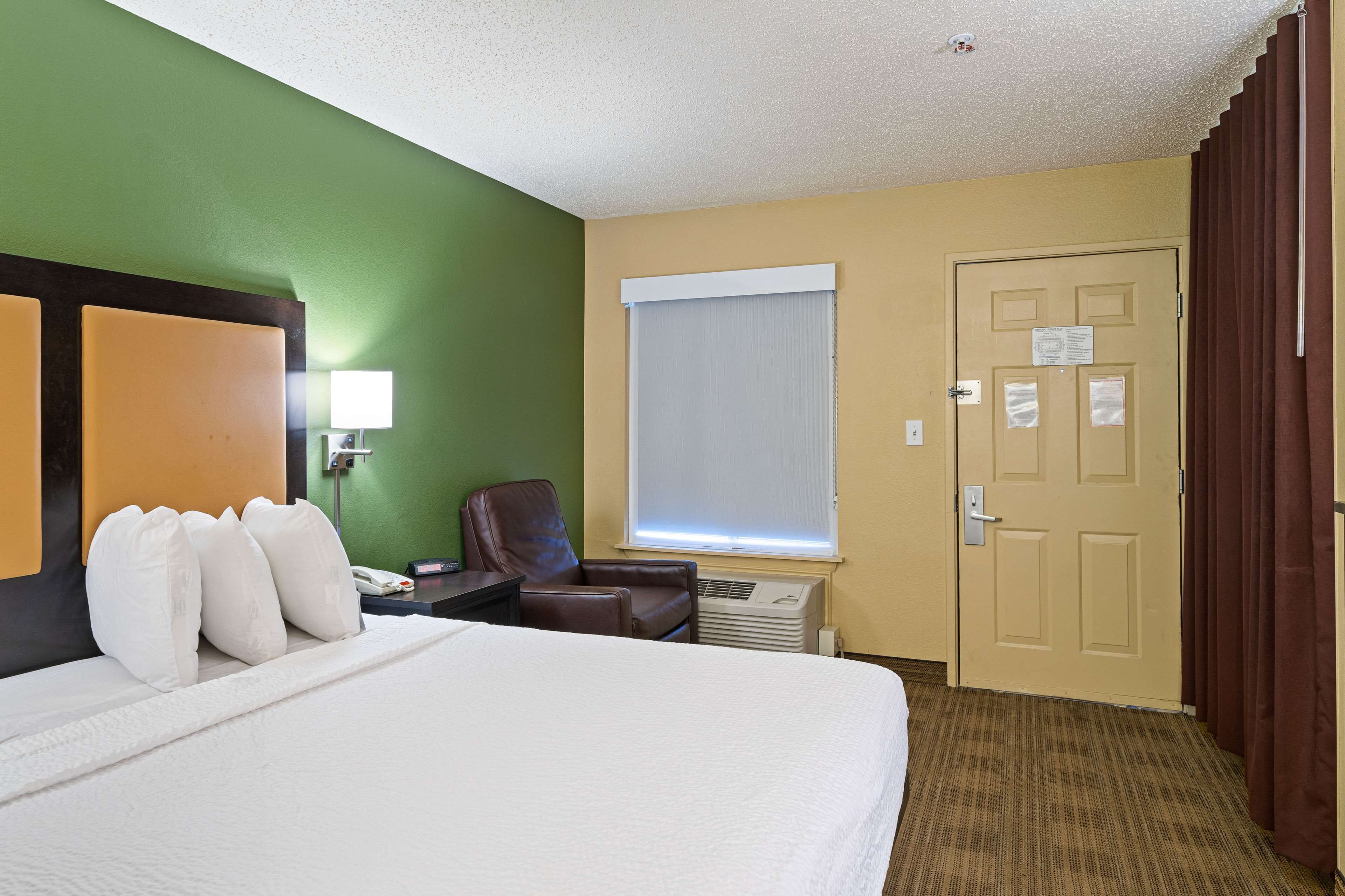 A hotel room with a bed, a chair, a desk with a lamp, a window, and a door.