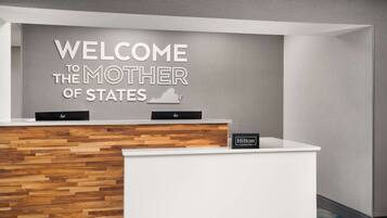 En receptionsdisk med et skilt der siger "Welcome to the Mother of States", træpaneldesign og et "Hilton Honors" badge.