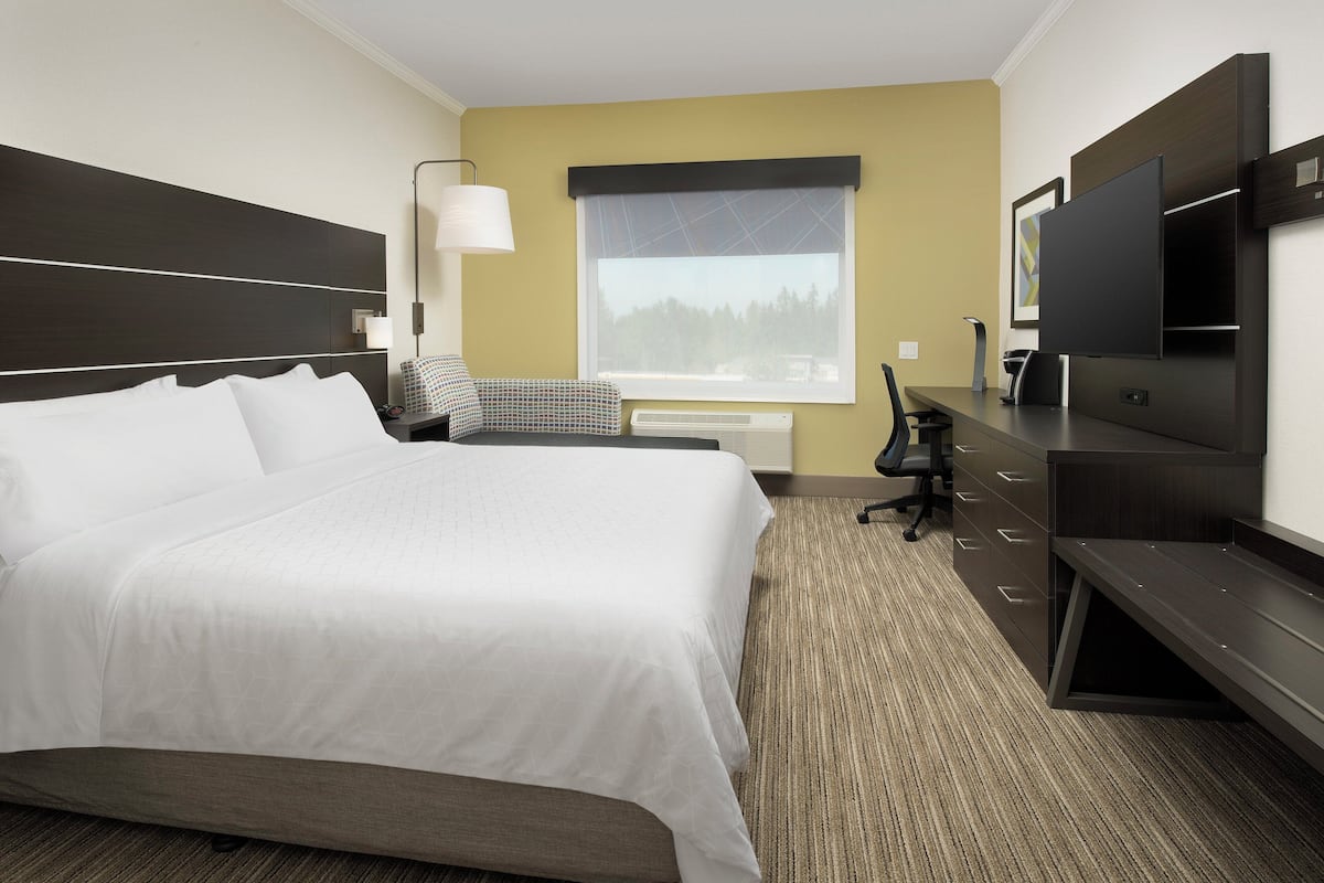 A hotel room with a large bed, a desk with a chair, a flat-screen TV, and a window with a view of trees.