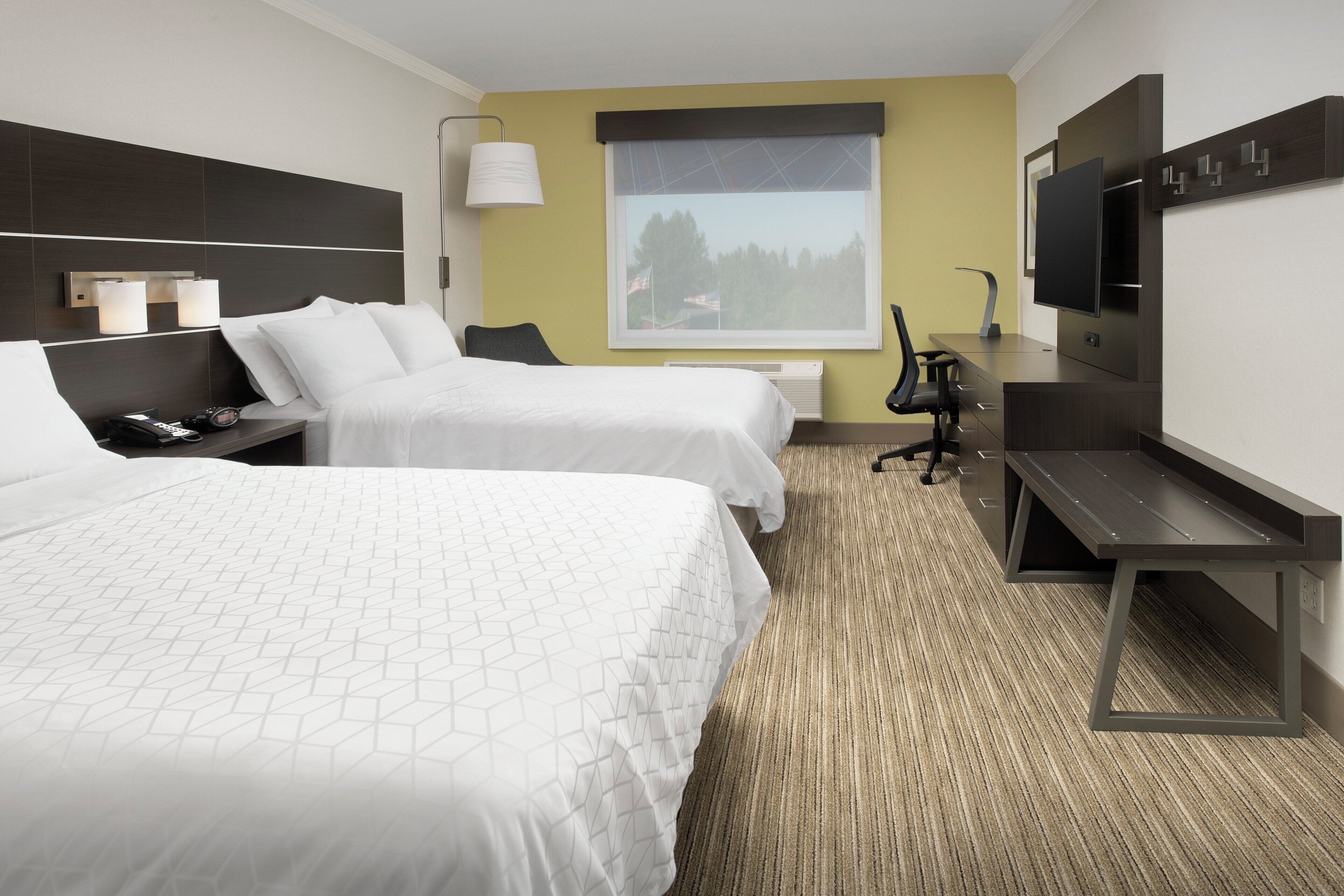 Ein Hotelzimmer mit zwei Betten, einem Schreibtisch, einem Stuhl, einem Fernseher und einem Fenster mit Blick auf Bäume.