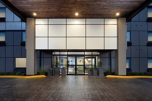 Una entrada de edificio moderna con techo de madera, ventanas amplias y zona pavimentada con jardineras.