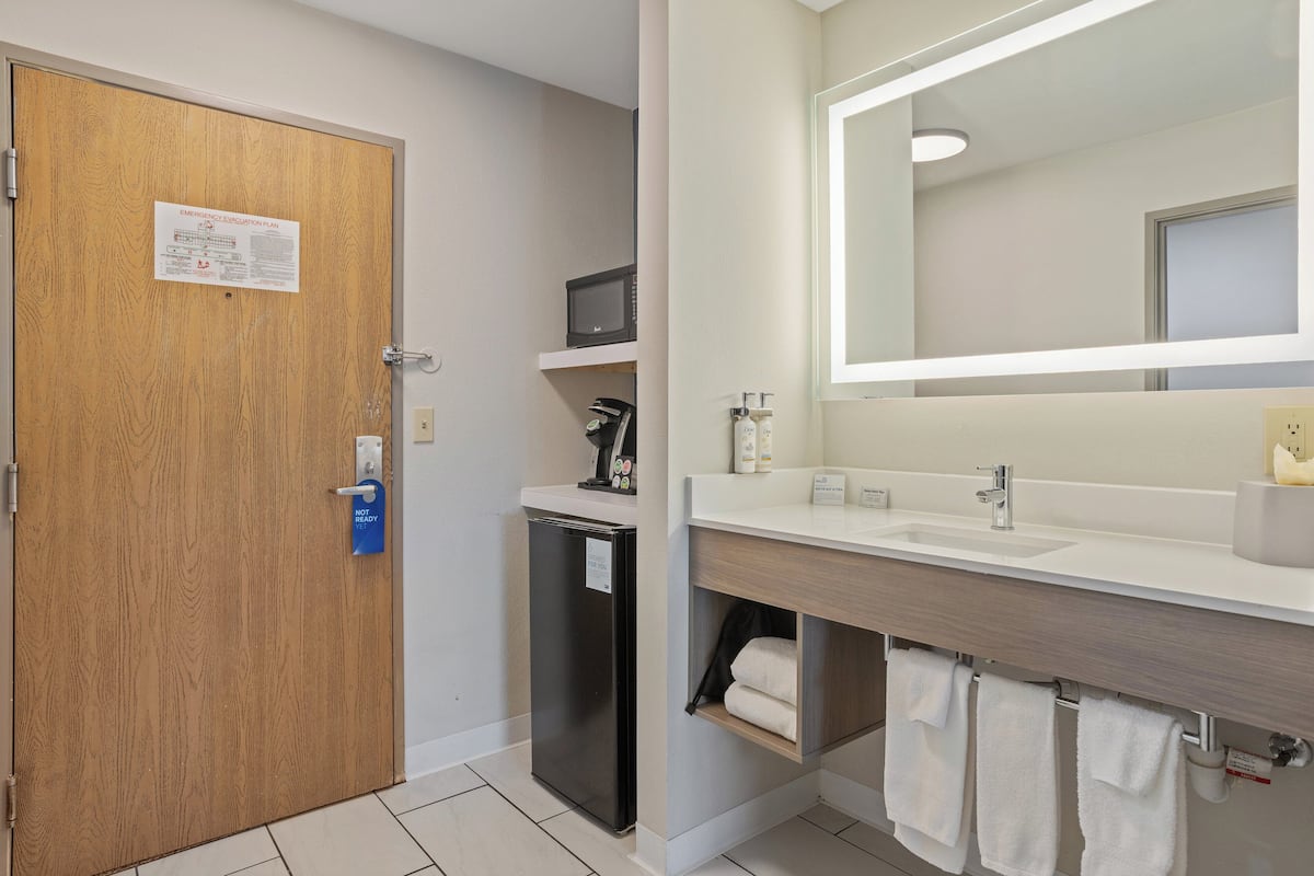 Un baño de hotel con puerta de madera, refrigerador, microondas, lavamanos y espejo.